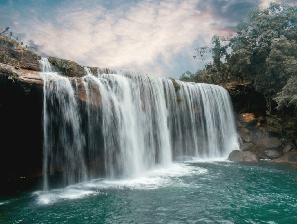 Peaceful waterfall cascading over rocks with lush forest backdrop and serene sky.